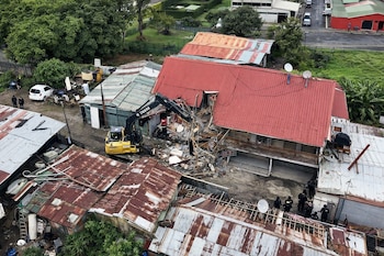 Una excavadora amarilla derriba un edificio de techo rojo, con escombros en el suelo, rodeado por otras edificaciones de chapa, personas y árboles.