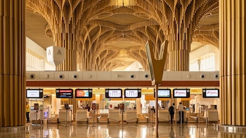 Vista interior de un aeropuerto moderno con mostradores de facturación, columnas acanaladas y un techo elaborado con listones de madera clara en forma de arco