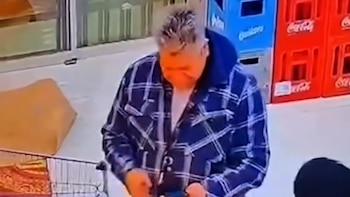 Hombre de cabello gris con camisa a cuadros azul y blanca, sudadera azul, manipulando un teléfono móvil junto a un carro de compras y cajas