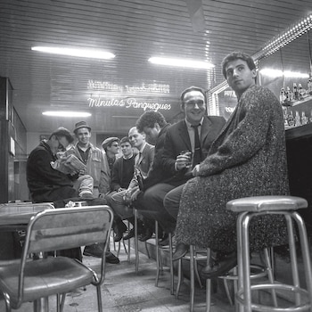 Fotografía en blanco y negro de varios hombres sentados en taburetes y sillas dentro de un bar con un letrero luminoso de fondo