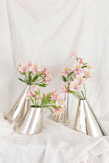 Cuatro floreros metálicos cónicos de color plateado contienen flores rosadas y blancas sobre una tela blanca arrugada