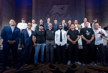Un grupo de hombres y una mujer posan de pie en dos filas frente a una pared con el logotipo de K-1 MMA sobre un escenario