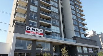 Edificio alto de color gris y blanco con múltiples pisos, balcones de vidrio y un gran cartel rojo y blanco en la fachada que dice "SE VENDE".