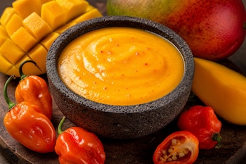 Cuenco de piedra con salsa amarilla espesa, rodeado de chiles habaneros anaranjados y rojos, y trozos de mango cortado.