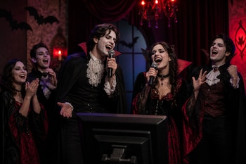 Cinco personas con disfraces y maquillaje de vampiro cantan con micrófonos frente a una pantalla en una habitación con decoración gótica.