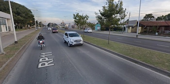 Una avenida de asfalto por donde circulan una camioneta utilitaria blanca y una motocicleta, junto a una franja de césped central y viviendas
