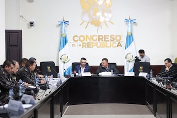 Personas con vestimenta formal conversan alrededor de una mesa con micrófonos y una bandera de Guatemala al fondo