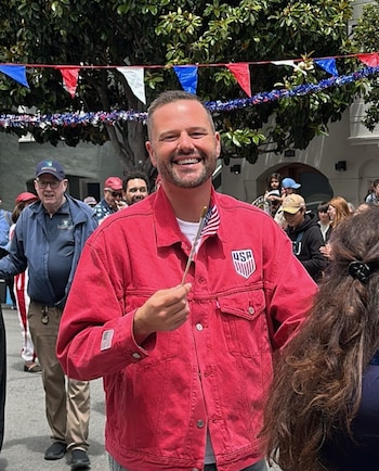 Hombre sonriente con barba y chaqueta roja que lleva insignias de Estados Unidos. Sostiene una bandera pequeña del país. Al fondo hay guirnaldas y gente