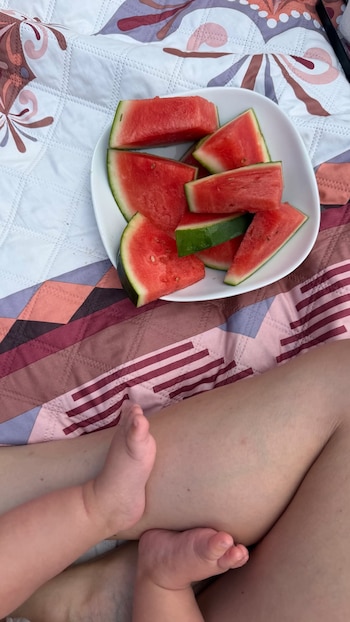 Un plato blanco con varios trozos de sandía de pulpa roja se encuentra sobre dos mantas estampadas. Los pies de un bebé reposan sobre la pierna de un adulto