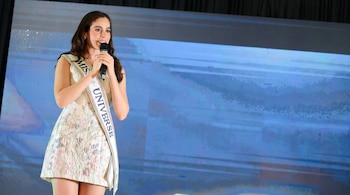Mujer con vestido estampado y banda de Miss Universe habla por micrófono ante una pantalla.