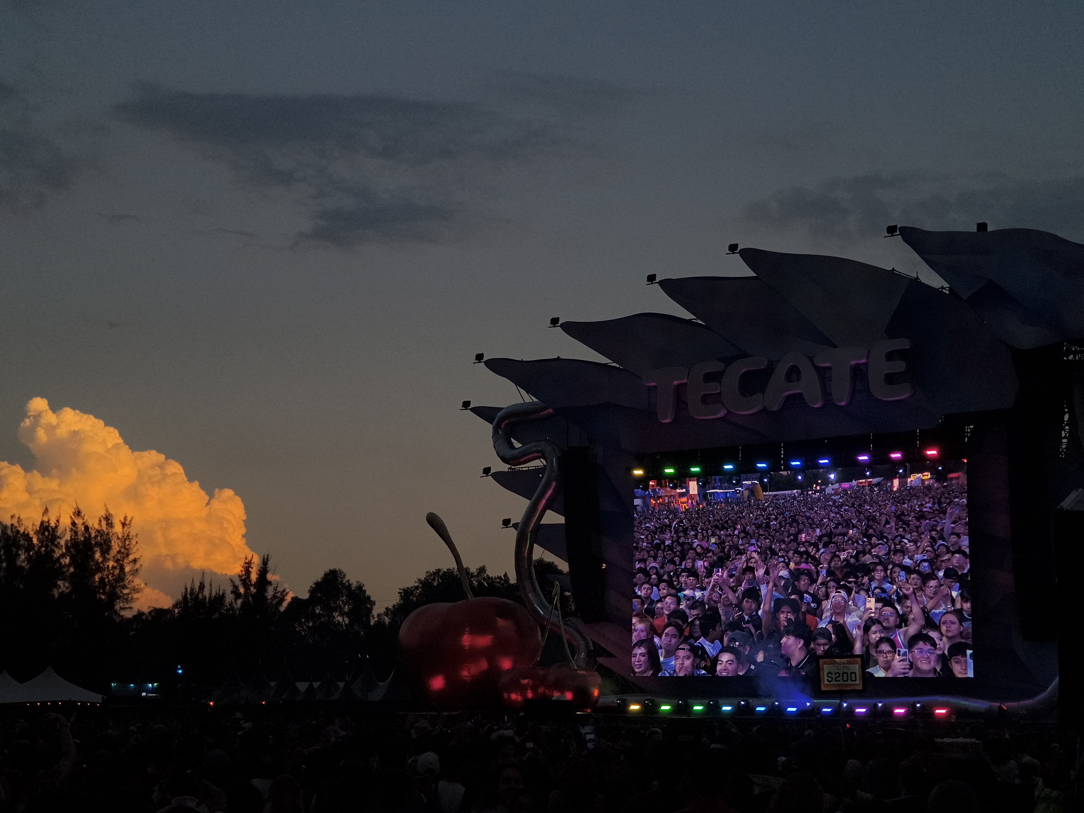 Miles de asistentes vibran frente al Tecate Emblema 2026, con la multitud reflejada en la pantalla gigante y una nube naranja iluminando el cielo al atardecer. (Carlos Adrian Ibarra)