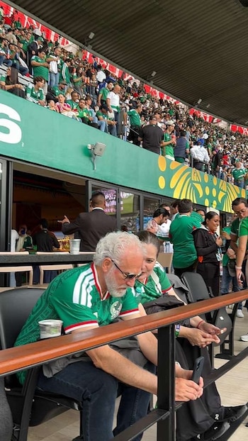Hombre con barba blanca y mujer con camiseta de México sentados en estadio; multitud de aficionados con ropa verde y blanca en gradas al fondo
