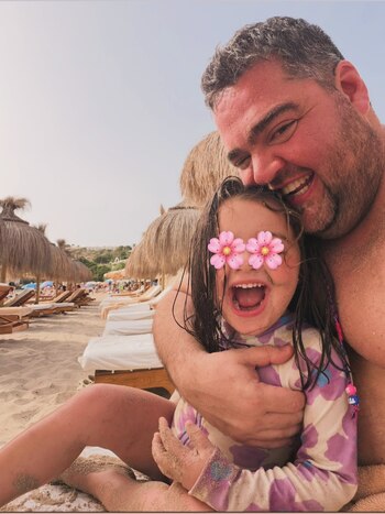 Un hombre y una niña ríen en primer plano, con arena en el rostro. La niña tiene flores digitales sobre los ojos. Detrás se observan sombrillas de paja y reposeras en una playa