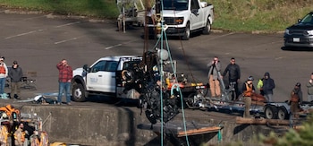 Una grúa levanta un coche oscuro y sumergido de una masa de agua, mientras personal de rescate y espectadores observan desde una rampa de hormigón