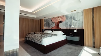 Dormitorio moderno con cama grande, ropa de cama blanca y manta de piel sintética. La pared tiene un mural abstracto y paneles de madera clara. Mesita de noche gris flotante