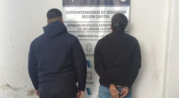 Vista trasera de dos hombres, uno con chaqueta oscura y otro con esposas, frente a un cartel de la Policía de Seguridad La Plata