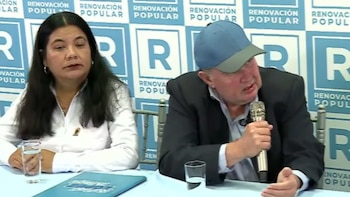 Rafael López Aliaga habla con un micrófono junto a una mujer frente a un panel publicitario del partido Renovación Popular
