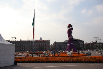 Figura gigante de una catrina con vestido morado rodeada por arreglos de flores anaranjadas y la bandera nacional en una plaza pública