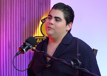 Un hombre con camisa oscura habla frente a un micrófono en un estudio decorado con paneles de madera