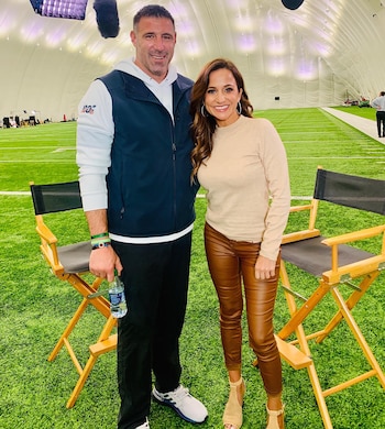 Mike Vrabel y Dianna Russini sonríen a la cámara de pie en un campo de fútbol artificial cubierto, con sillas de director de madera detrás de ellos