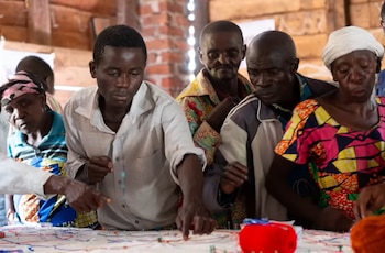 Un grupo de adultos, incluyendo hombres y mujeres, se inclina sobre una mesa, señalando un mapa o diagrama con numerosos alfileres de colores brillantes