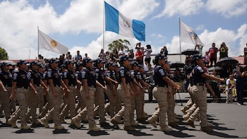 Grupo de policías con uniforme azul marino y caqui, gorras y parches de la bandera de Guatemala, marchando. Al fondo, espectadores, árboles y dos banderas