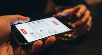 Primer plano de una mano sosteniendo un móvil con una app de citas abierta y un "match" difuso; en segundo plano, otra mano sostiene un vaso borroso.