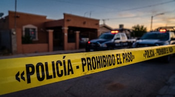 Una cinta amarilla de prohibición policial en primer plano con patrullas y una casa al fondo.