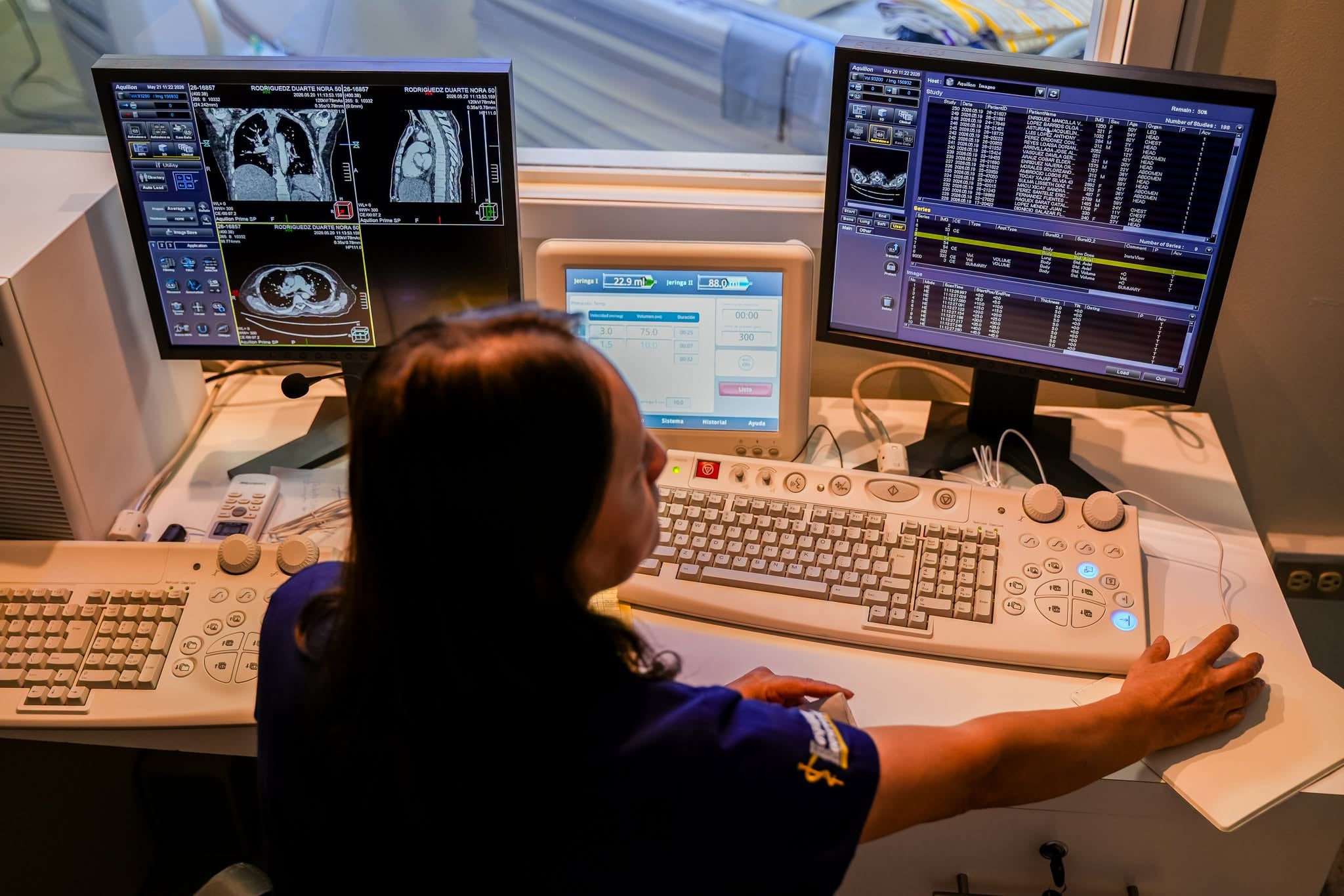 Una profesional de la salud opera una consola de tomografía computarizada con pantallas que exhiben escaneos médicos y listas de pacientes. (Ministerio de Salud y Asistencia Social)