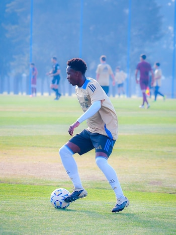 Un joven futbolista negro con camiseta dorada y azul, pantalones cortos oscuros, medias blancas con logo AFA y botines negros conduce un balón en un campo de césped