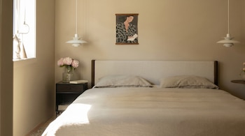 Dormitorio con cama doble, ropa de cama beige, cabecero tapizado, mesita de noche con jarrón de peonías y libro. Pared color arena con cuadro y luz solar.