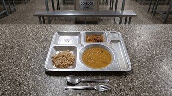 Una bandeja metálica con arroz, sopa y guiso sobre una mesa de granito junto a una cuchara, un tenedor y bancas al fondo.