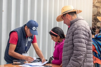 Un hombre con gorra azul y chaleco atiende a una mujer con suéter rosa y a un hombre con sombrero de paja en una mesa con documentos