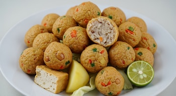 Un plato blanco repleto de bolitas de batagor doradas. Una de ellas está cortada, revelando su relleno. Incluye trozos de tofu, patata y medio limón verde.