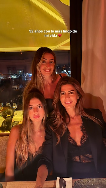 Juliana Awada y sus dos hijas posan en un restaurante con iluminación cálida, frente a una ventana con luces de la ciudad de fondo. Las tres sonríen