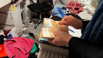 Manos contando billetes de 50 euros. De fondo, una habitación desordenada con ropa. En la esquina superior izquierda, el logo de la Guardia Civil