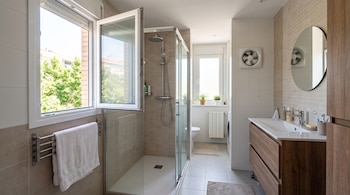 Un baño iluminado con ventana abierta, mampara de ducha de cristal, lavabo de madera, espejo redondo y un extractor en la pared.