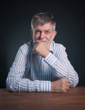 Retrato de un hombre de mediana edad con cabello y barba grises, camisa a rayas azules y blancas, sonriendo mientras apoya su barbilla en la mano