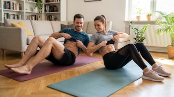 Un hombre y una mujer sentados en colchonetas de yoga en una sala de estar, sonríen mientras levantan sus camisetas para mostrar abdominales tonificados.