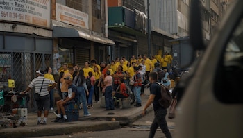 Grupo de personas en una calle, varias con camisetas amarillas y chalecos. Edificios, letreros de tiendas, acera y un vehículo parcialmente visible