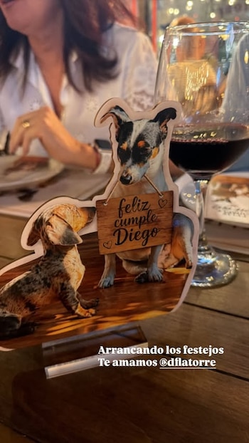 Dos figuras de perros recortadas sobre una mesa de madera. Un perro sostiene un cartel que dice "feliz cumple Diego". Una copa de vino tinto y una persona borrosa