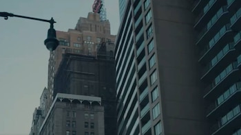 Un rascacielos clásico y un moderno edificio de apartamentos de gran altura se erigen contra un cielo gris, con una farola en primer plano