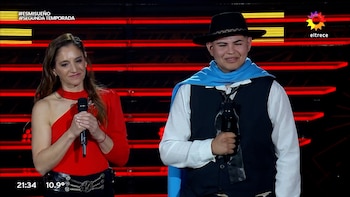 Una mujer con blusa roja y un hombre con sombrero, poncho azul y chaleco sostienen micrófonos frente a un fondo con luces rojas
