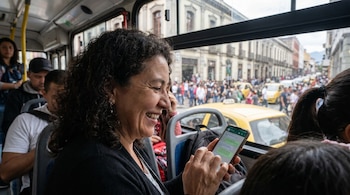 Mujer sonriente con cabello rizado oscuro sentada en un autobús, usando su teléfono móvil. Vista de una calle urbana concurrida con peatones y taxis amarillos.