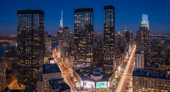 Vista aérea nocturna de Manhattan con rascacielos altos, edificios con luces encendidas y apagadas, y trazas de luz de vehículos en las calles debajo.