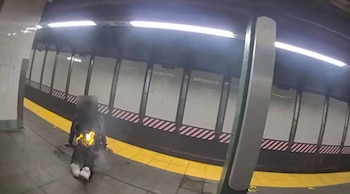 Una persona sentada en un andén de metro tiene fuego en la espalda, con una franja de seguridad amarilla en el suelo y el túnel de vías al fondo