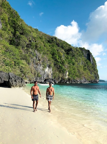 Dos hombres caminan descalzos por una playa de arena clara junto a aguas turquesas, con grandes acantilados rocosos cubiertos de vegetación al fondo