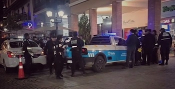 Varios policías y dos vehículos policiales, uno tipo sedan y otro camioneta, con luces azules encendidas, se encuentran en una calle de noche junto a un cono naranja