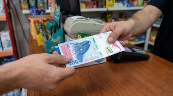 Dos manos sostienen un billete de 200 pesos argentinos sobre un mostrador junto a una caja registradora y un terminal de cobro.