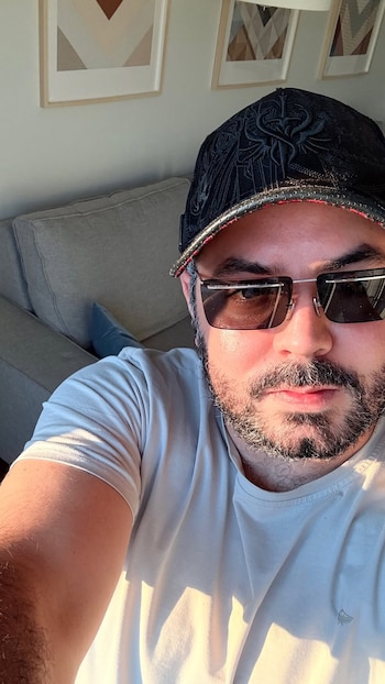 Hombre con barba y bigote usa gorra negra y gafas de sol, camiseta blanca; al fondo, sillón gris y cuadros en la pared con figuras geométricas